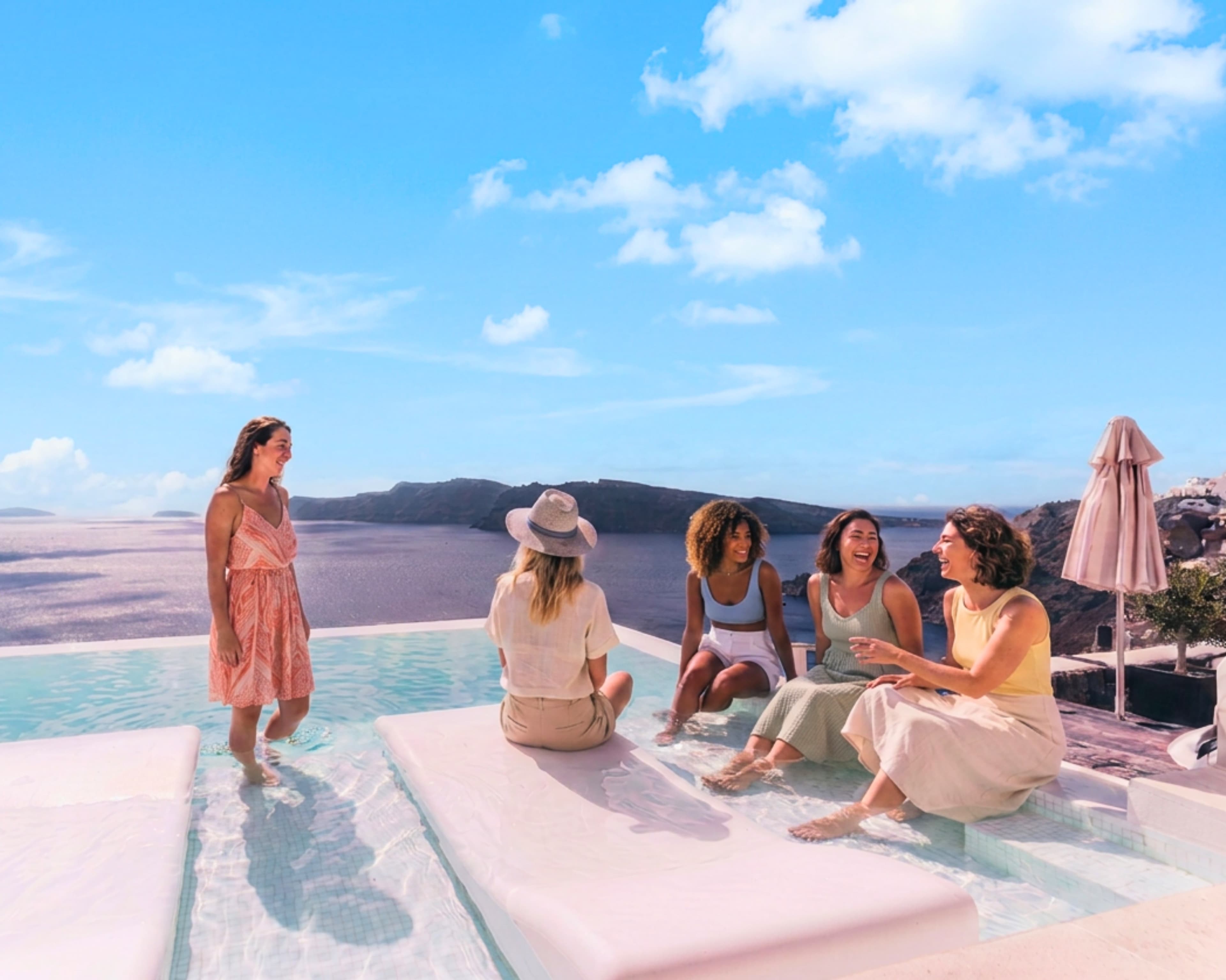 Five women laughing together at the edge of an infinity pool above the Santorini caldera