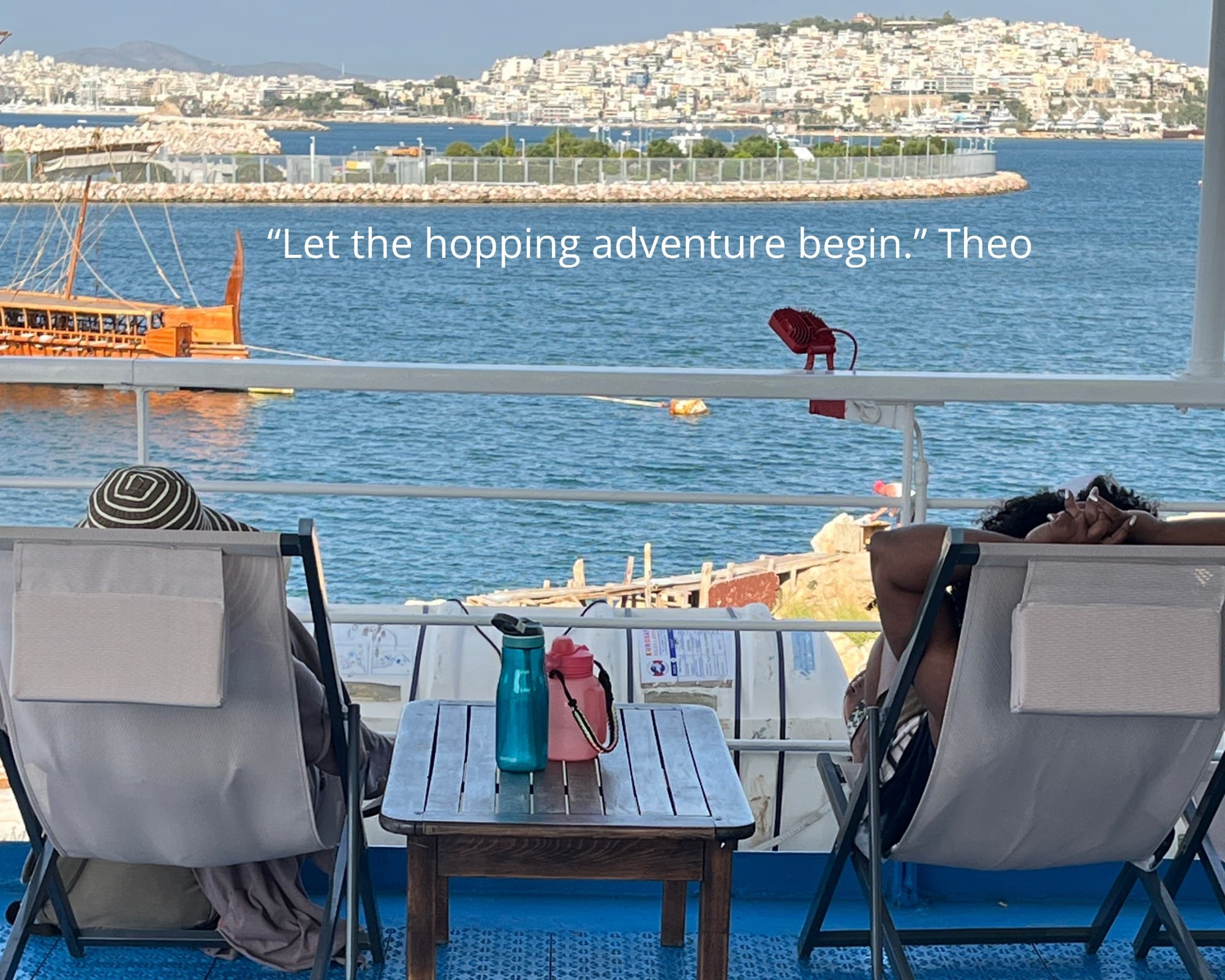 Two travellers in deck chairs on a boat, looking across the water to the town on the hill - Monobrow Greek island hopping