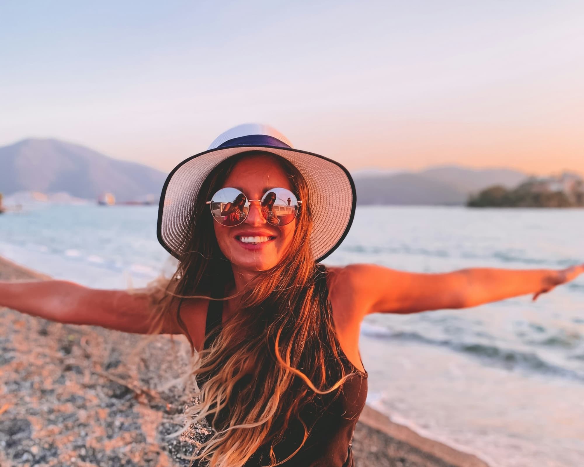 A traveller in a sun hat standing at the water's edge on a Greek beach at sunset, arms outstretched - Monobrow Holidays Greece